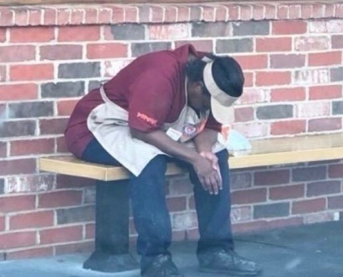 A worker in a maroon Popeyes uniform slumps over on a bench with his head down in exhaustion, perfectly capturing the despair of desperately hoping for a cryptocurrency moonshot to escape the daily grind. The Popeyes logo on his apron contrasts humorously with his prayer for XRP to surge to $589.