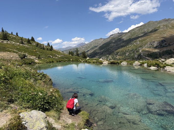 A scenic mountain landscape featuring a clear turquoise alpine lake, with a person in a red backpack sitting at the water's edge surrounded by rocky peaks and green vegetation.