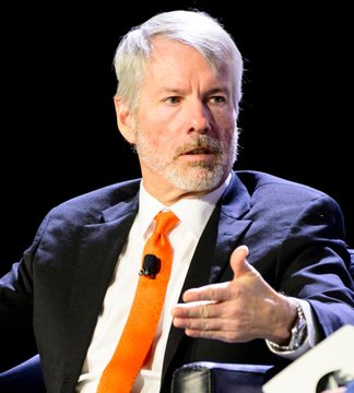 A middle-aged man with white hair and a beard, wearing a dark suit with a bright orange tie, gestures with his hand while speaking, with a microphone clipped to his lapel.