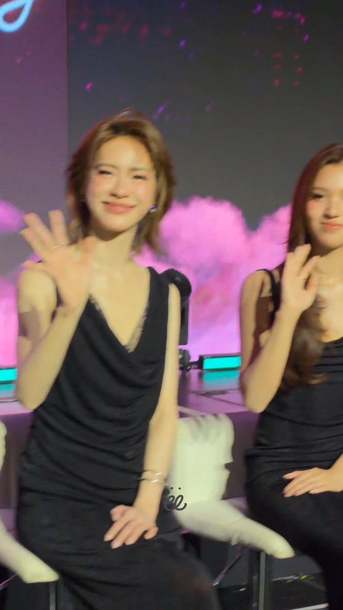 A woman in a black cowl-neck dress waves to the camera while smiling, seated next to another woman at an event with pink smoke effects in the background.