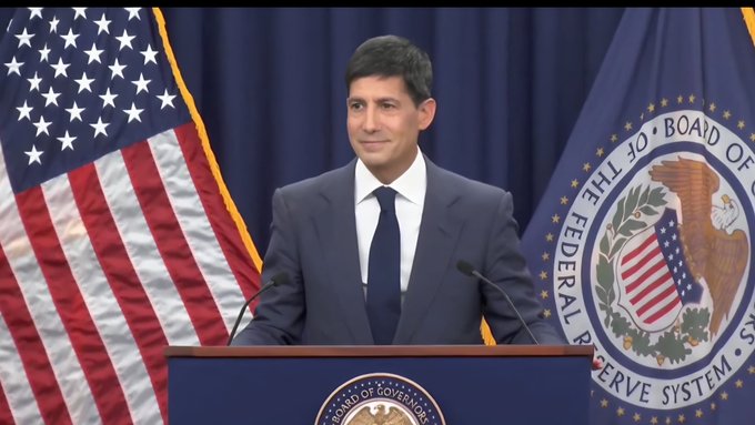 A man in a dark suit stands at a podium between the American flag and the seal of the Federal Reserve Board of Governors.