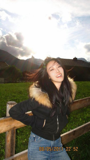 A casual photo of a smiling young woman wearing a black jacket with a fur-lined hood, standing by a wooden fence in a grassy field with mountains in the background, timestamped '08/05/2011 22:52'.
