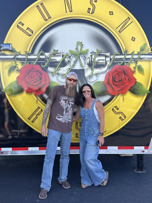 A man with a long beard wearing sunglasses and a graphic t-shirt standing next to a woman in denim overalls and sunglasses, posing in front of the Guns N' Roses tour bus logo.