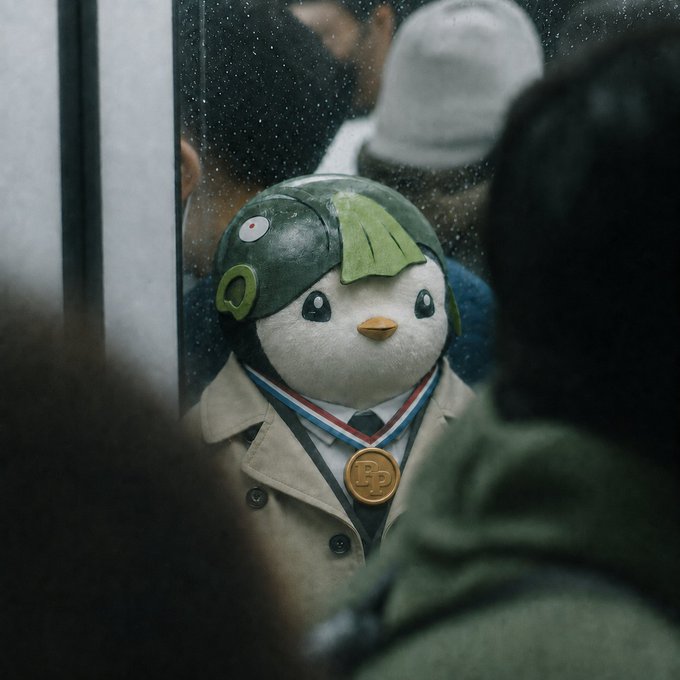 A plush penguin toy wearing a green helmet and beige trench coat with a gold 'B' medal around its neck, displayed behind glass with a person visible in the foreground.