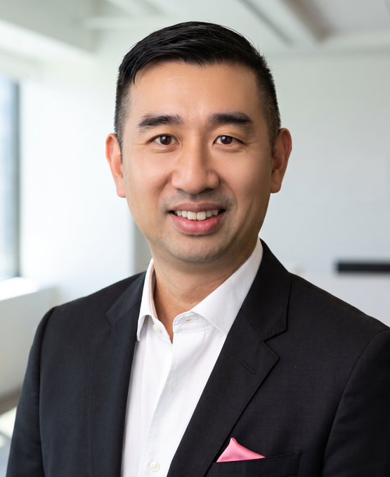 A professional headshot of a smiling East Asian man wearing a dark gray suit jacket, a white collared shirt, and a pink pocket square, photographed against a bright, out-of-focus background.