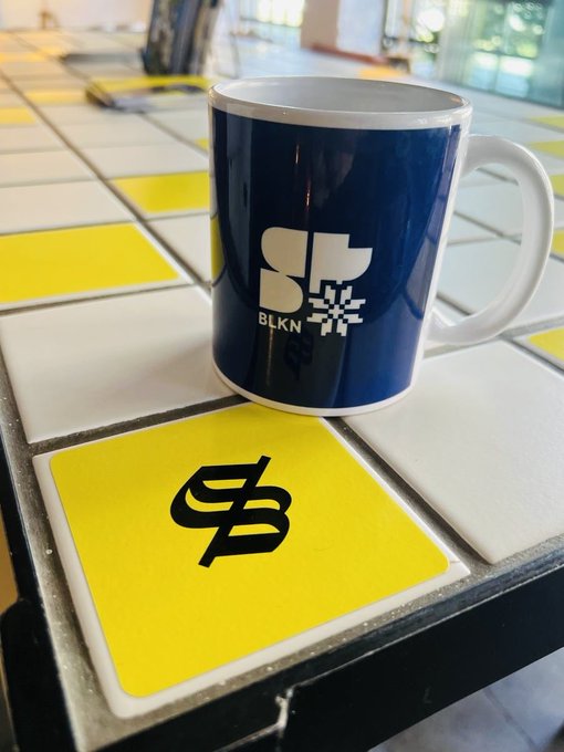 A blue ceramic mug displaying the 'BLKN' logo sits on a tiled table next to a yellow tile featuring a black stylized 'B' currency symbol.