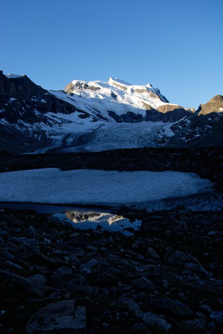 A scenic landscape featuring snow-capped mountains under a clear blue sky, with a glacier and a small reflective pool of water in the rocky foreground.