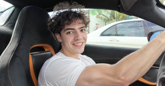 A young man with curly brown hair wearing a white t-shirt sits in the driver's seat of a sports car with black and orange interior, smiling at the camera.