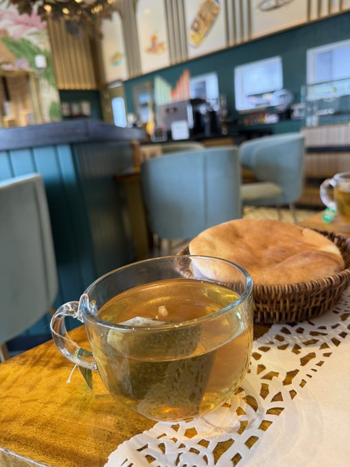 A close-up of a clear glass mug filled with light tea with a tea bag, sitting on a wooden table next to a basket of bread in what appears to be a cafe setting with teal chairs and decor.