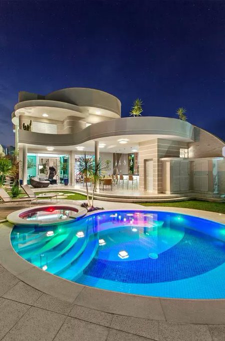 A luxurious modern curved home at night features a stunning curved swimming pool illuminated with vibrant blue, green, and pink lights, complete with a circular spa, lounge chairs, and palm trees under a starry sky.