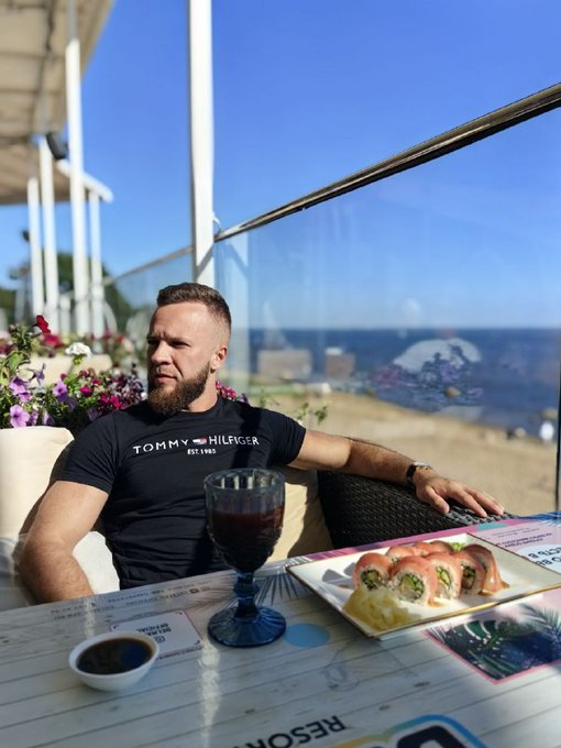 A bearded man in a black Tommy Hilfiger t-shirt sits at an outdoor seaside restaurant table with sushi, a glass of red wine, and ocean views.