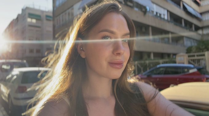 Close-up selfie of a woman with long brown hair outdoors in sunlight, with buildings and parked cars in the background.