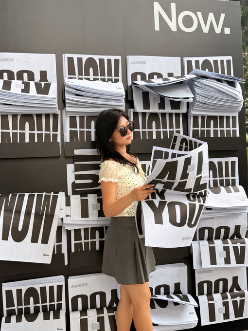 A young woman in sunglasses, polka dot top, and gray skirt holds up large posters spelling 'WOW' and 'YOU' in front of a wall covered in stacks of posters and 'Now.' text.