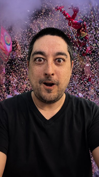 A selfie-style photo of a surprised-looking man in a black v-neck shirt, with a surreal background of pink and purple confetti and floating objects.