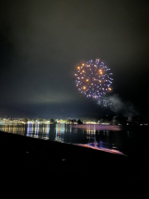 Fireworks bursting in colorful patterns over a calm body of water at night, with lights from a distant shoreline reflecting on the surface.