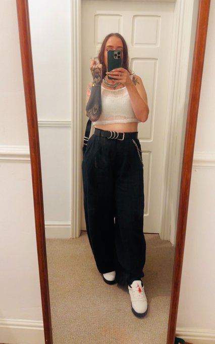 A mirror selfie shows a person wearing a white crop top and black wide-leg pants with a belt, standing in front of a white door.