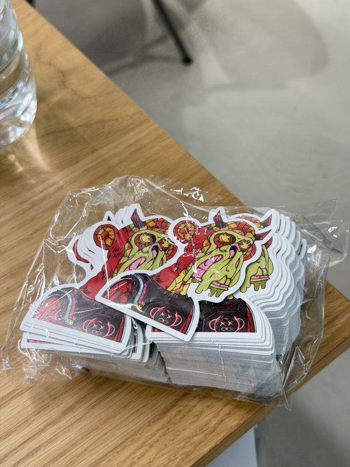 A sealed plastic bag of die-cut stickers featuring a horned, fanged ape mascot in red and yellow tones sits on a wooden table, ready to be claimed by the next holder of the associated NFT collection.
