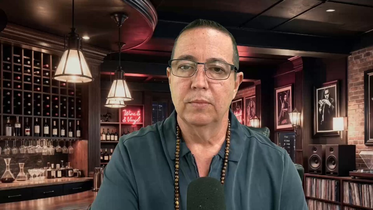 The image features a man wearing glasses, sitting at a bar with several bottles of wine behind him. The man appears to be talking into a microphone or recording device, possibly discussing something related to the wine selection available at the bar. The background includes a large picture and some lights that create an atmosphere for socializing and enjoying drinks.
