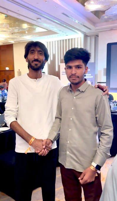 Two men shake hands at the Binance event in Islamabad. One man is wearing a white shirt and black pants, while the other is wearing a grey shirt and maroon pants.