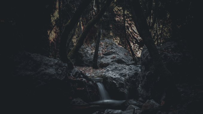 A moody, dark photograph of a small waterfall in a rocky forest gorge, with smooth long-exposure water flowing through shadowy trees and boulders.
