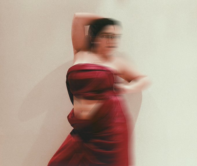 A motion-blurred photograph of a woman with dark hair wearing a flowing red dress, posing dynamically against a plain light-colored background.