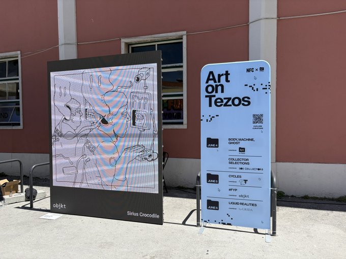 The image features a large poster with black text that says "Art on Tezos" and "Two curations. Two moods." The poster also includes an image of a map, which could be related to the theme or context of the art exhibit. The background of the image is pink, possibly indicating the location where the exhibit is taking place. There are two smaller posters in the scene as well, one located near the center and another towards the right side.
