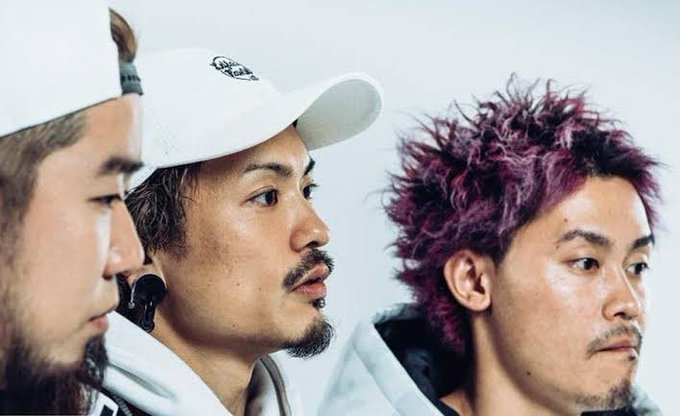 Three men are pictured in a candid close-up, with the man in the center wearing a white cap featuring the text "City Boys," while the man on the right sports vibrant purple hair—appearing to be a group photo from the JASMY/HARA project team's social media. The image seems to represent the team behind JASMY, whose CFO Hara is tagged in the post discussing whether JASMY is a permissioned consortium chain and why it's been placed on Binance's monitoring zone.