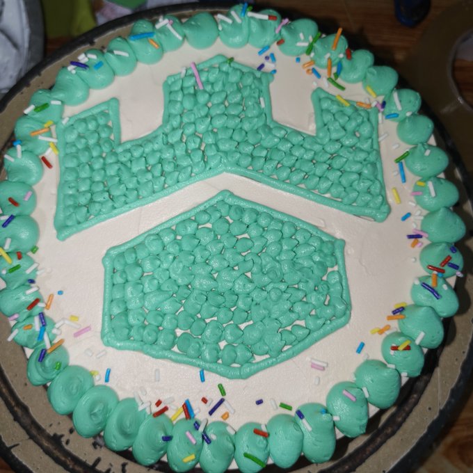 A round cake featuring green frosting decorated to resemble the Gamdom logo, complete with a crown design and colorful sprinkles around the border, was made by a fan as a tribute to SteveWillDoIt's $HERO token and Gamdom partnership.