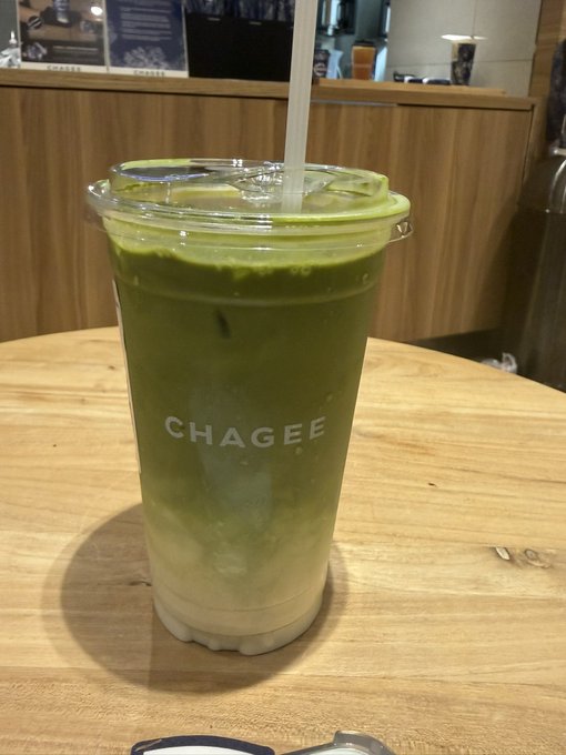The image shows a cup of green tea sitting on top of a wooden table, with a straw sticking out of it. The cup has the word "Chaegee" written on it, indicating that this is a specialty drink from a Japanese cafe or restaurant. The background features a dining area and some other cups, suggesting that this might be a popular spot for enjoying tea in a cozy atmosphere.