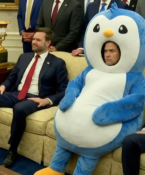 Vice President JD Vance sits beside a person in a blue penguin mascot costume, with other suited officials visible in the background.