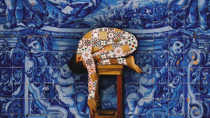A person draped over a wooden stool wearing a patterned outfit, posed against a wall of traditional blue and white azulejo tiles.
