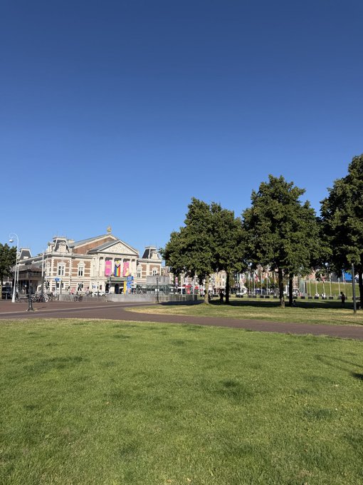The Concertgebouw, Amsterdam's iconic concert hall, stands prominently under a brilliant blue sky on a sunny Sunday, with its grand classical facade visible across the open green of Museumplein.