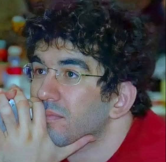 A close-up portrait of a man with curly dark hair and glasses, wearing a red shirt and resting his chin on his hands in a thoughtful pose.