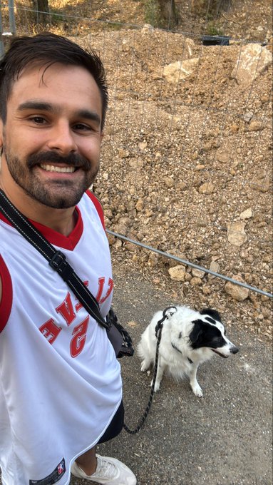 A smiling man in a red and white sleeveless shirt takes a selfie with a black and white border collie dog on a leash, with a rocky, dirt-covered hillside in the background.