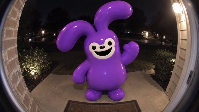 A large purple inflatable bunny character with a smiling face stands on a doorstep at night, viewed through a fisheye lens, greeting visitors.