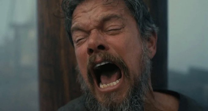 A bearded man with his eyes closed and mouth wide open screams in apparent anguish, set against a misty, grayish background.