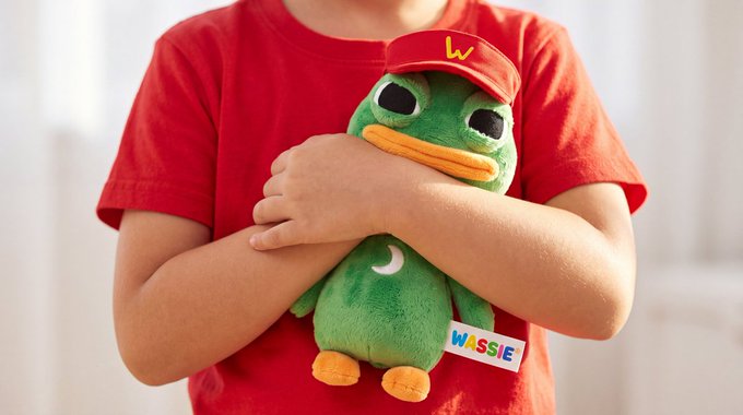 A child in a red shirt cradles a green duck plushie wearing a red "W" cap, with a tag reading "WASSIE." The image playfully illustrates the post's message, suggesting the cute Wassie mascot is what's preventing viewers from getting their own small Wassie plushies.