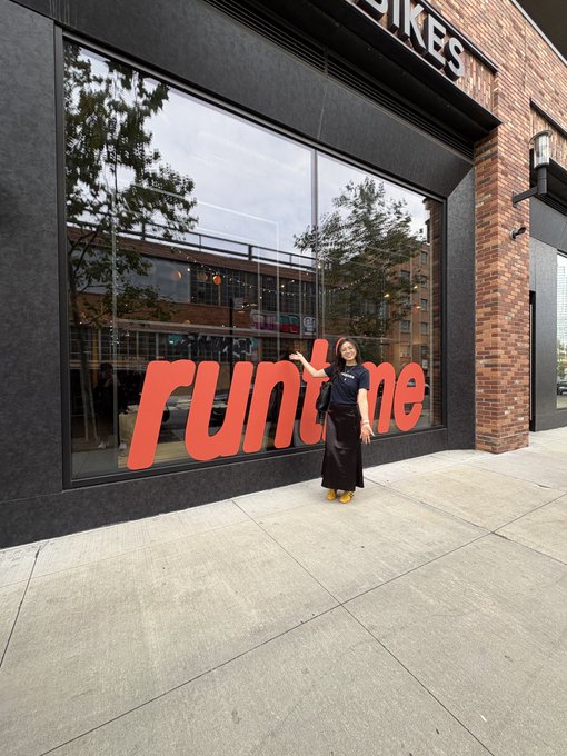 A photo of a woman posing in front of a storefront window with a large red 'runtime' sign, with part of a sign above reading 'IKES'.