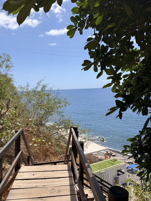 A wooden staircase descends toward a sunny coastal cove with clear blue waters and a small beach, framed by lush green foliage. The image captures the essence of the "60 minutes" prompt—suggesting a quick escape to a secluded seaside spot before getting back to daily life.