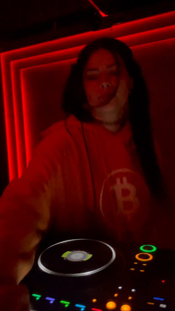 The image features a woman wearing an orange sweatshirt with a white B logo, standing next to a DJ mixer or turntable. The background of the photo has red neon lights that create a vibrant atmosphere. There are also some text elements in the image, including "@sautrah playing heaters at bass night in a Bitcoin sweater" and "@sautrah this one goes out to the Bitcoin $DOG army".