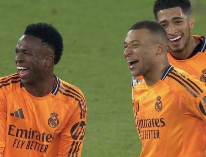 Real Madrid players including Vinícius Júnior, Kylian Mbappé, and Jude Bellingham, wearing the orange 'Emirates FLY BETTER' away kit, smile and laugh together on the pitch.