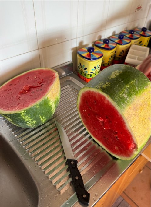 A kitchen sink scene with two halved watermelons and a black-handled knife on the drainboard, with decorative yellow ceramic canisters in the background.