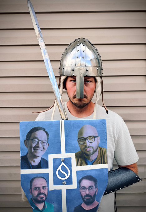 A person stands wearing a medieval metal helmet with a nose guard, holding a sword and a blue shield painted with four men's faces around a central logo.