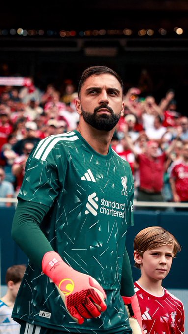 Liverpool goalkeeper Alisson Becker stands on the pitch wearing his green goalkeeper jersey with "Standard Chartered" sponsorship, alongside a young fan in a red kit.

This image fits the "Good Morning ft" caption by showcasing Alisson, likely before a match, providing a bright, fan-filled start to the day.