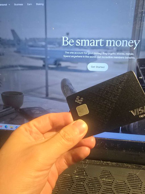 A hand holding a black Visa card in front of a laptop displaying a website with the headline 'Be smart money,' describing 'The one account for your money. Buy crypto, stocks, metals. Spend anywhere in the world. Get incredible members benefits.'