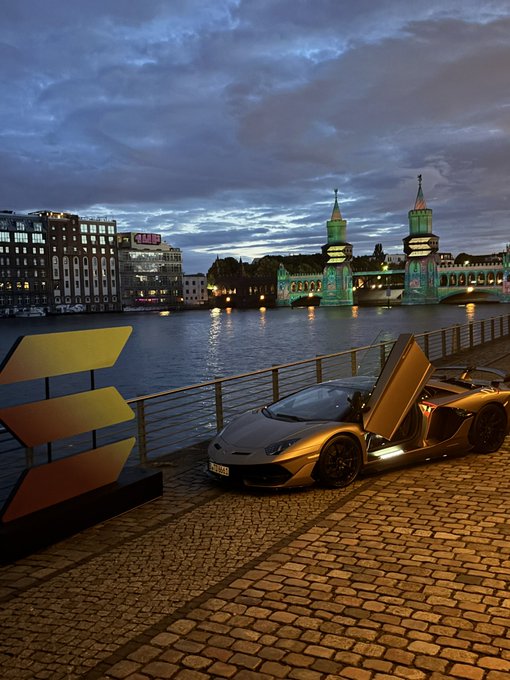 The image features a silver sports car parked next to a brick walkway by a body of water with two bridges in the background. The car has its trunk open, revealing an interior view. A sign can be seen nearby, possibly indicating parking regulations or directions for visitors.