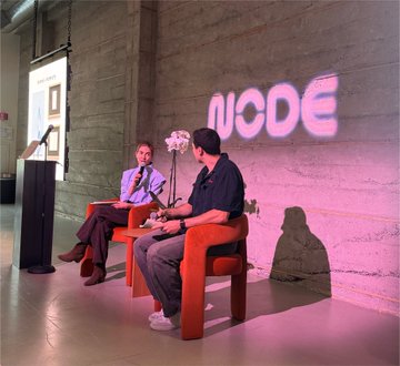 Two men seated in orange chairs on a stage in an industrial-style venue with pink neon "NCODE" signage on a concrete wall behind them, appearing to be at a fireside chat or panel discussion.
