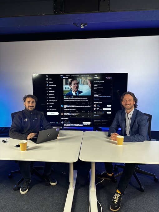 The image shows two men sitting at a table with laptops open in front of them. They are positioned so that they can have a video call or participate in an online meeting, possibly discussing current market sentiment or Bitcoin yield. The room has a blue wall behind them and a large TV screen mounted on the wall. There is also a cup placed on one of the tables between the two men.