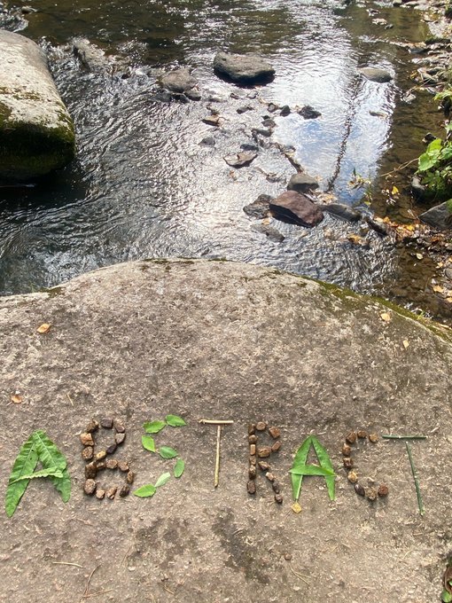 The word "ABSTRAIT" is spelled out using natural materials—leaves, twigs, and acorns—on a large flat rock next to a shallow stream.