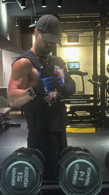 A muscular man wearing a black cap, black tank top, and blue workout gloves looks at his smartphone in a gym, with two '15 KG HAMMER STRENGTH' dumbbells resting in the foreground.